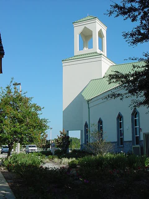 Exterior view of a white church with green roofing and a bell tower surrounded by greenery and a walkway with a parking lot
