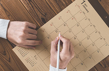 A top-down view of a person’s hands using a pencil to mark a date on a tan-coloured paper wall calendar sitting on a wooden desk.
