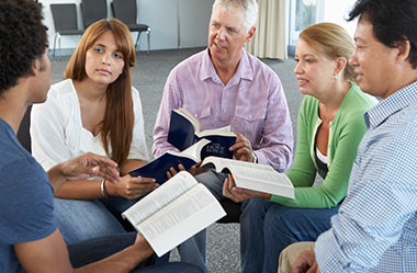 A diverse group of five adults of various ages sit in a circle in a brightly lit room, holding open Bibles and engaging in a thoughtful group discussion.