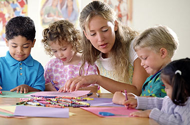 A smiling female teacher or volunteer leans over a table to help a diverse group of elementary-aged children with an art project involving colorful construction paper, crayons, and safety scissors.